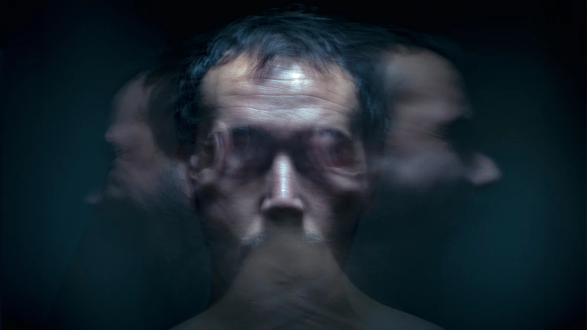 A dark closeup of a man's face shrouded in shadow. Other blurry and transparent versions of the face can be seen laughing on each side of him implying his insanity.